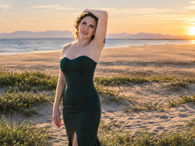 TessaDaybreak, met krullend bruin haar, staat op een zandstrand in een strakke groene jurk zonder bandjes, haar hand in haar haar, waarbij ze haar borsten en een sensuele glimlach laat zien..