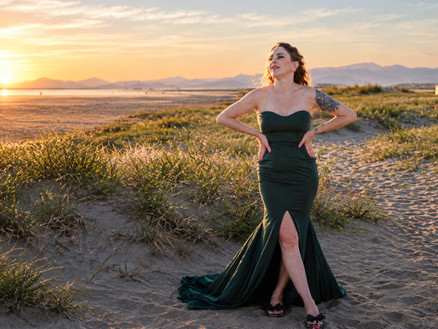 TessaDaybreak steht an einem sandigen Strand, trägt ein grünes, trägerloses Kleid mit einem hohen Schlitz, hat die Hände in die Hüften gestemmt und ein Tattoo am rechten Arm. Im Hintergrund ist ein Sonnenuntergang zu sehen..