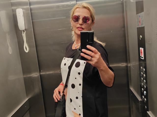 SexyCoco taking a selfie in an elevator, wearing pink reflective sunglasses, black shirt with white polka dots, black bag, blonde hair in a ponytail, holding a phone.