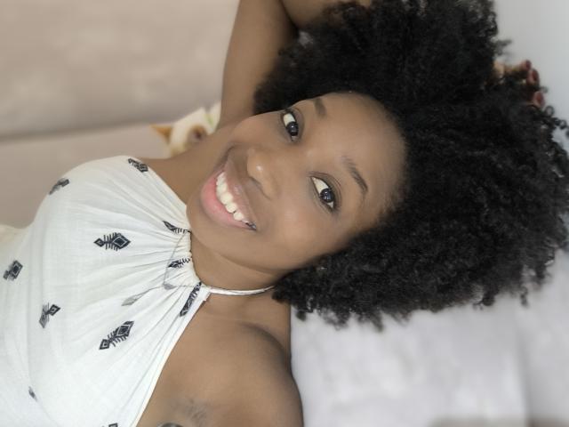 XMeryVegaXi with dark skin, curly black hair, smiling, wearing a white halter top with black diamond patterns, arms raised, showing armpit hair, lying on a bed.