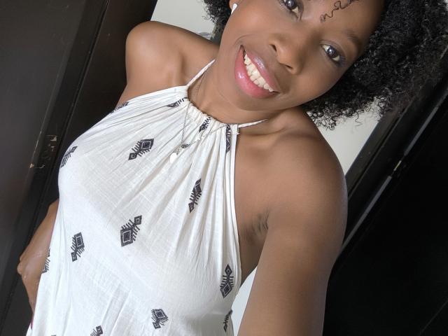 XMeryVegaXi with curly black hair, smiling, wearing a white halter dress with black leaf patterns, showing her tits, armpit, and a small tattoo above her right eyebrow.