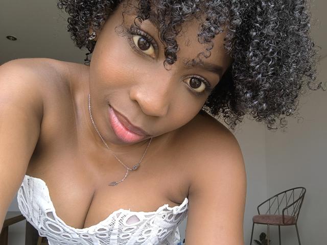 XMeryVegaXi with curly black hair, brown eyes, and a white lace strapless top, showing her tits, smiling at the camera, wearing a silver necklace, in a selfie.