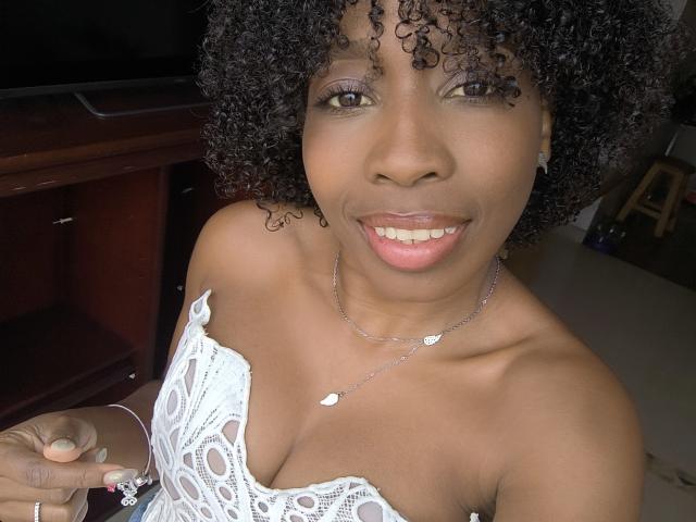 XMeryVegaXi with curly black hair, smiling, wearing a white lace strapless top, showing her tits, holding a ring, silver necklace, and earrings, in a selfie.