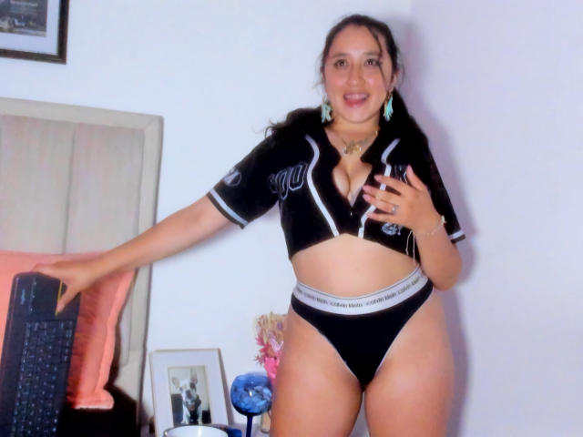 JessyBarnnett with dark hair in pigtails, wearing a black cropped shirt and black Calvin Klein panties, hand on chest, blue earrings, smiling, standing in front of a TV.