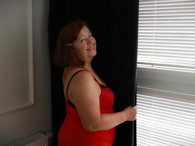 AdeleLoveEx con un vestito rosso, reggiseno nero e capelli castani corti, sorridente, che tiene una tenda nera, in piedi vicino a persiane bianche, mostrando i suoi fianchi prosperosi e i suoi piccoli seni..