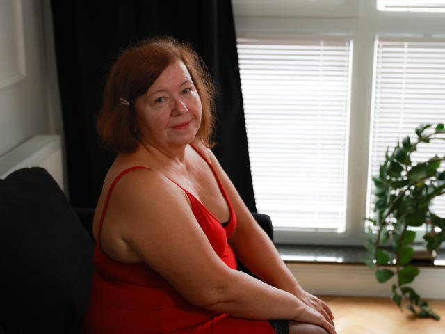 AdeleLoveEx tiene el pelo rojo, un cuerpo regordete y pechos pequeños. Lleva un vestido rojo, está sentada en un sofá negro y sonríe a la cámara. Se pueden ver sus brazos..