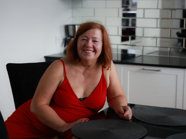AdeleLoveEx, une rousse corpulente avec de petites seins, sourit dans une robe rouge, assise à une table noire avec des assiettes grises, dans une cuisine moderne..