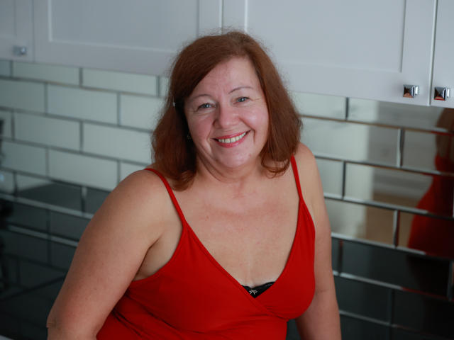 AdeleLoveEx, a chubby redhead woman with medium tits in a red tank top, smiling, sits in a kitchen with white tiles and cabinets. Her skin is fair.