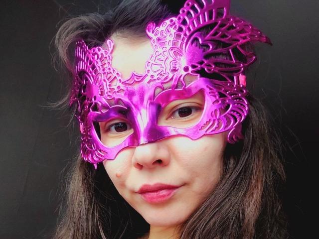 IrieBirdy wearing a shiny purple mask with intricate patterns, has brown wavy hair, and a slight smile with pink lipstick, in a dark background, looking at the camera.