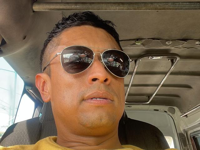 Hachesah has tan skin, short black hair, and is wearing aviator sunglasses with a reflection of a car's interior. He's in a yellow shirt, looking slightly to the side.