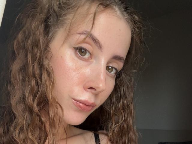 EmiliaSue has curly brown hair, green eyes, and shiny skin with a slight blush. She's looking at the camera with a subtle smile, wearing a black strap.