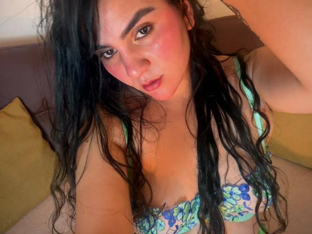 FenixAlthea with long, wavy black hair, pink cheeks, and green-blue floral bra, looking at the camera with a slightly open mouth, showing her tits. Soft lighting.