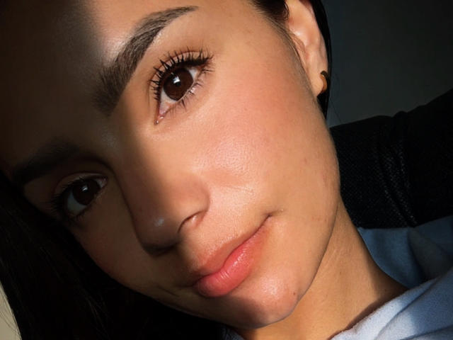 KimmyK's face, brown eyes, slightly parted pink lips, light brown skin, dark eyebrows, small black ear studs, dark hair, white shirt, soft light on face, subtle smile.