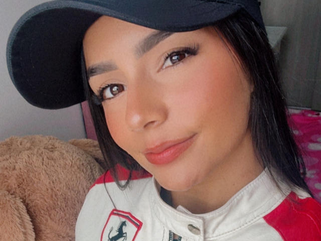 KimmyK with tan skin, black eyebrows, brown eyes, and long black hair, wearing a black cap and red-and-white racing shirt, smiling subtly, showing a cute, sexy face.
