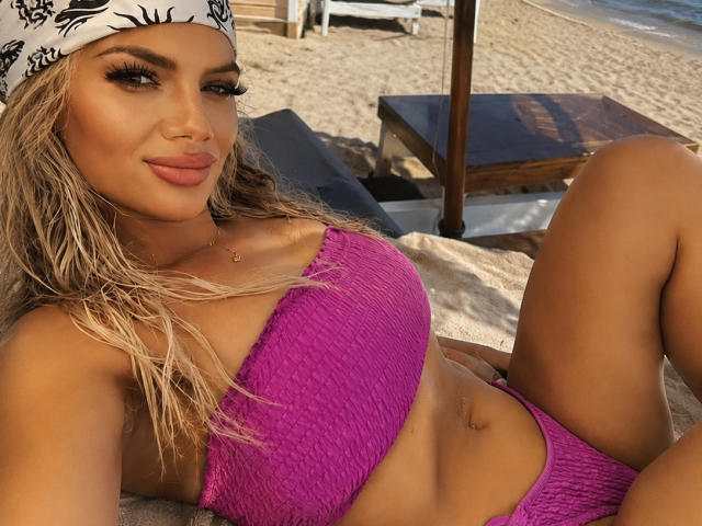 LisaAnnMarys mit blonden Haaren, weißem Stirnband und rosa Top und Hose, liegt am Strand, zeigt ihre Brüste und ihren kleinen Bauchnabel, lächelt mit vollen Lippen..