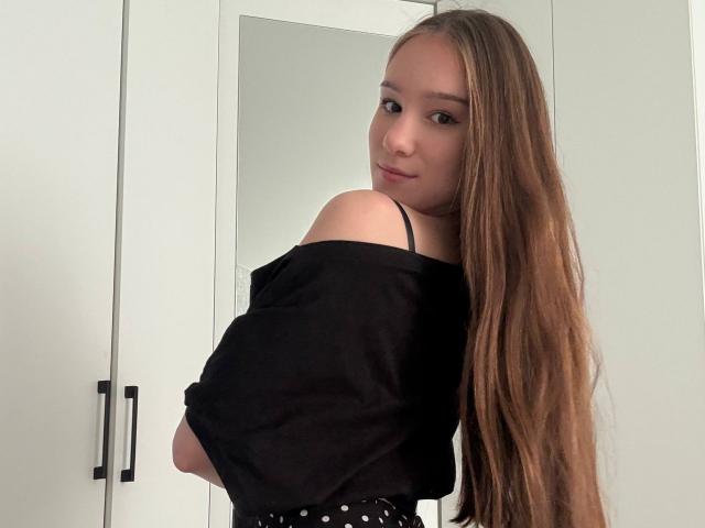 DomingaSierra met lang bruin haar, een zwarte top met een open schouder en een rok met stippen, kijkt om en glimlacht zachtjes, waarbij haar blote schouder en zwarte bandje zichtbaar zijn..