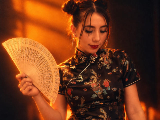 FreyaraVenwo with dark hair in double buns, wearing a shiny black floral cheongsam, holding a lace fan, looking down with red lipstick, in warm orange lighting.