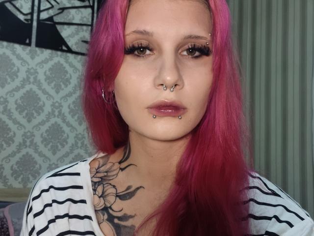ShenaBatrum, con cabello rosa, aro en la nariz, piercings en los labios y un tatuaje en el pecho, viste una camisa a rayas, tiene pestañas largas y ojos verdes. Su trasero no es visible..