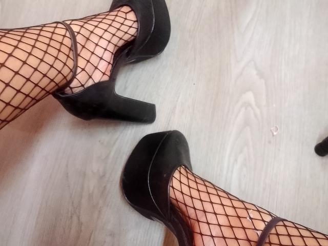 Le gambe di Chloee Williams, con calze a rete nere e tacchi alti, mostrano i suoi piedi su un pavimento di legno. La sua vulva non è visibile..