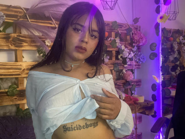 ChiaraBella with long brown hair, wearing a white off-shoulder top, exposing her titty and "Quibibog" tattoo on her boob. Purple light in the background. Slightly pouty expression.