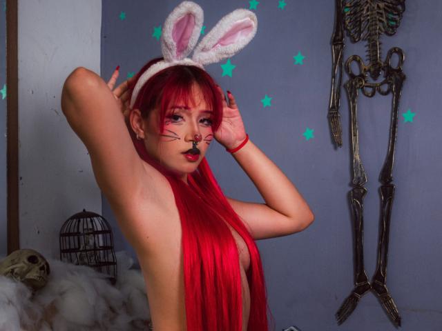 Chloee Williams, con cabello rojo, orejas de conejo blancas, maquillaje en la cara y cabello rojo largo, sin ropa, con las manos detrás de la cabeza, mostrando los pechos, en una habitación espeluznante con decoración de esqueletos..
