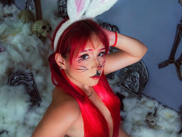 Chloee Williams, con cabello rojo, orejas de conejo y maquillaje negro, tiene un busto grande, lápiz labial rojo y un piercing en la nariz, y está de pie en una sustancia blanca y esponjosa, con calaveras y piezas de metal..