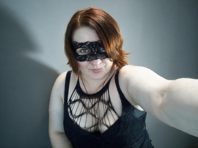 AgilaMipo with red hair, wearing a black lace mask and black lace-up top, exposing her tits, holding a selfie stick, smiling at the camera.