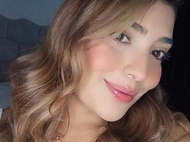 BiancaRey tem cabelos castanhos ondulados e brilhantes, lábios rosados e um sorriso adorável, usando um colar dourado, com bochechas levemente coradas e olhos castanhos olhando para a câmera..