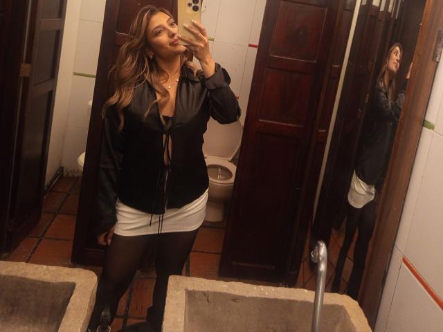 BiancaRey, avec de longs cheveux bruns, une veste noire, une mini-jupe blanche, des collants noirs et des bottes, prend un selfie devant un miroir, montrant ses seins et ses fesses dans une salle de bain..