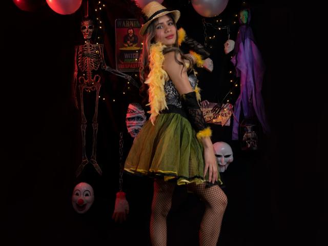 Sheyla Spooner in een circusoutfit, met een gele verenboa, een groene rok, een zwarte kanten top, netbroek en een hoed, met skeletten en clownmaskers op de donkere achtergrond..