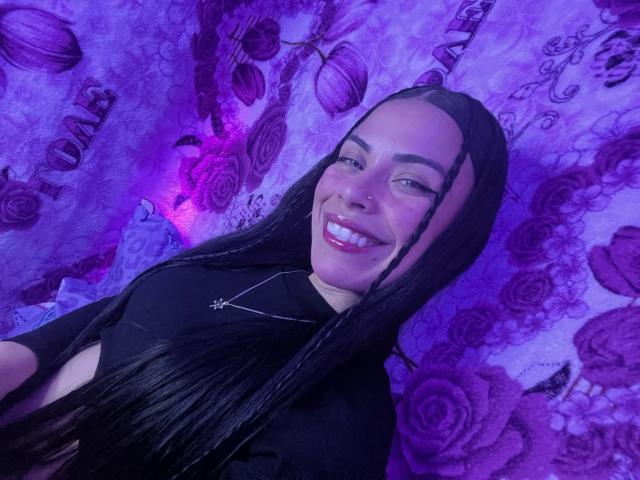 ZoeLux with long black hair, smiling, wearing a black top, and a cross necklace, has white skin and purple lighting, showing her teeth and looking at the camera.