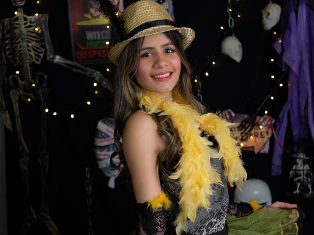 Sheyla Spooner, con una boa di piume gialle, guanti di pizzo nero, un cappello di paglia, sorridente, con i capelli castani, un corpetto grigio, che mostra il seno, in un allestimento a tema Halloween con scheletri e decorazioni spaventose..