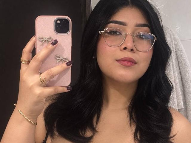 AlexaKiim mit schwarzen, welligen Haaren, Brille, pinkem Lippenstift und dunklen Nägeln, hält ein pinkes Telefon mit Schmetterlingsdekor, in einem Badezimmer-Selfie, auf dem ihr Gesicht und der obere Brustbereich zu sehen sind..