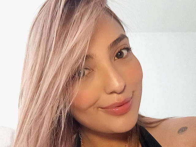 BiancaRey, avec ses cheveux bruns clairs et des mèches blondes, cligne de l'œil et sourit, montrant ses lèvres roses et brillantes, avec un petit tatouage sur l'épaule droite, portant un haut noir, peau claire, mignonne..