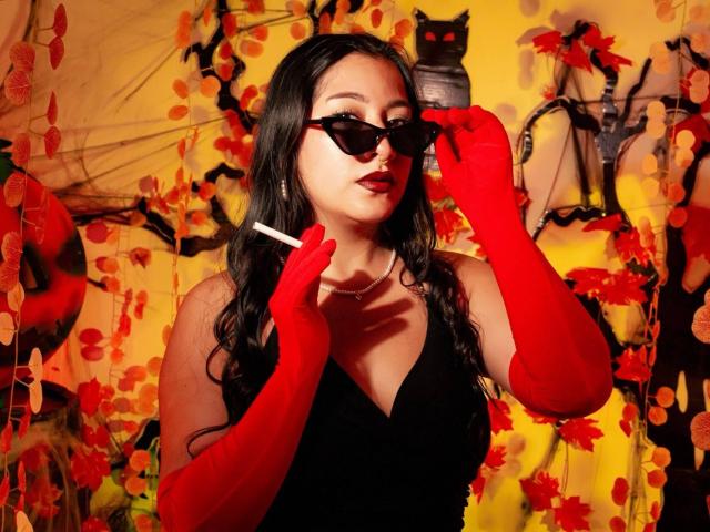 Paula Loughty, con cabello oscuro, usando guantes rojos y gafas de sol negras, sosteniendo un cigarrillo, con una blusa negra, y un fondo de hojas amarillas y naranjas..