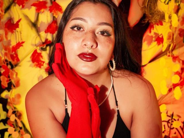 Paula Loughty, con cabello oscuro, lápiz labial rojo, piercing en la nariz y una blusa negra, sosteniendo una bufanda roja, con aretes dorados, fondo amarillo y rojo, mirando a la cámara, con expresión seria..