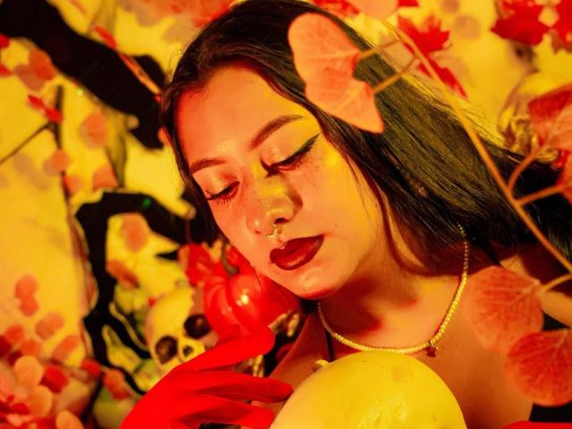 Paula Loughty, con capelli scuri, trucco occhi giallo, rossetto rosso, piercing al naso e una collana, che tiene foglie gialle e rosse, circondata da fiori rossi e teschi, in una luce gialla calda..