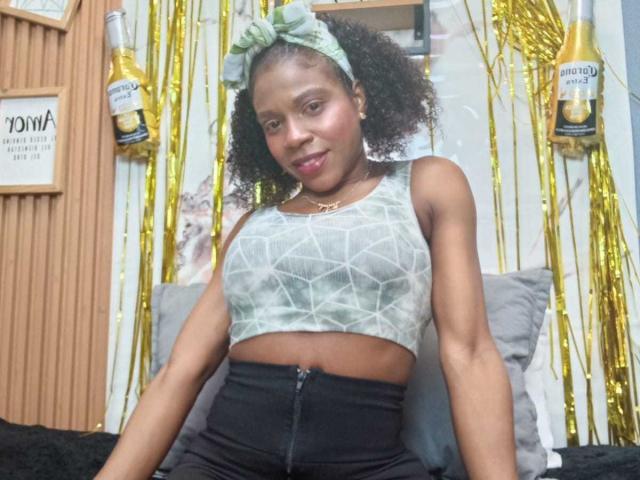 Sarahi Pusey, con cabello rizado, usando una diadema verde, un top corto gris de diseño geométrico, pantalones negros de cintura alta, sentada en una cama, sonriendo, con serpentinas doradas y botellas de cerveza en el fondo..