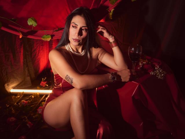 AfroditaDoir with dark hair, red dress, pearl necklace, tattoo on arm, sitting, crossed legs, wine glass, red and black lighting, seductive expression, wearing bracelet, red fabric background.