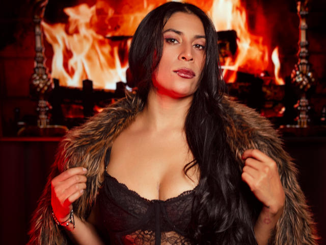 AfroditaDoir with long black hair, wearing a lace bra, furry coat, red bracelet, and red lipstick, standing in front of a roaring fire. Her tits are prominent.