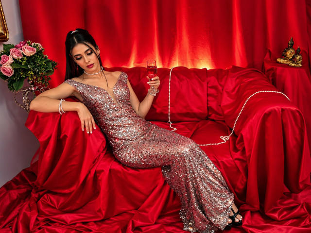 Anna Alvarez, vêtue d'une robe rouge scintillante ornée de sequins, est assise sur des meubles recouverts de satin rouge, tenant un cocktail rouge, ses talons noirs et son bracelet étant visibles, avec des roses roses à proximité..