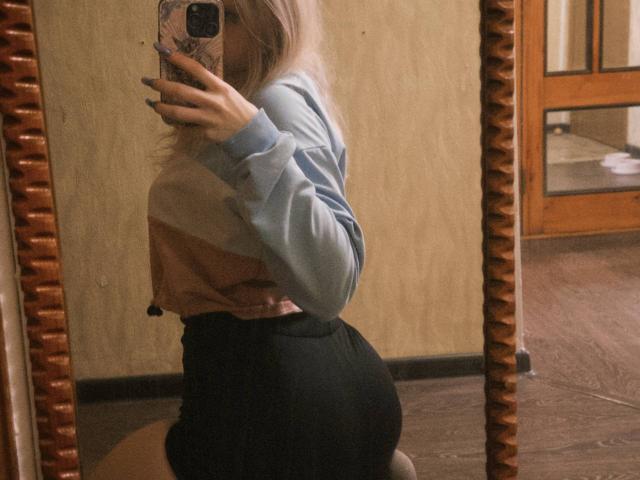 TiffaniMacandog dans un selfie pris dans un miroir, montrant ses fesses dans un short noir taille haute, portant un pull rayé rose et bleu, et tenant un téléphone avec une coque colorée..
