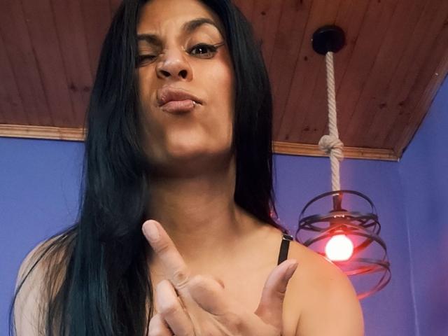 AfroditaDoir with long black hair, winking, and pouting, wearing a black top, pointing with her index finger at the camera, in a room with a wooden ceiling.