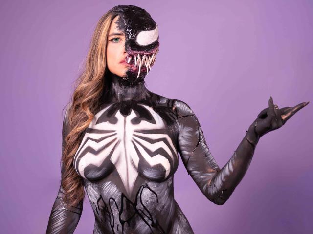 SofyaLane in a black Venom cosplay suit with white spider web chest, black gloves, and a Venom mask, pointing with her right hand, long blonde hair, purple background.