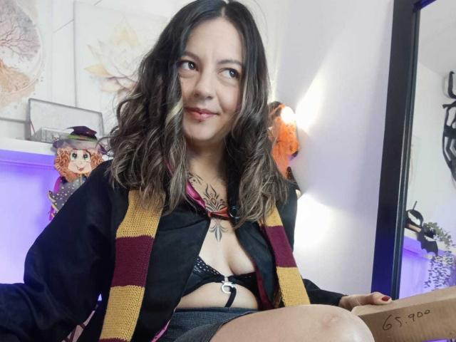 ArianaBlonde with wavy brown hair, smiling, wearing a black top with a burgundy and yellow striped scarf, black lace bra, and gray shorts, showing her tits.