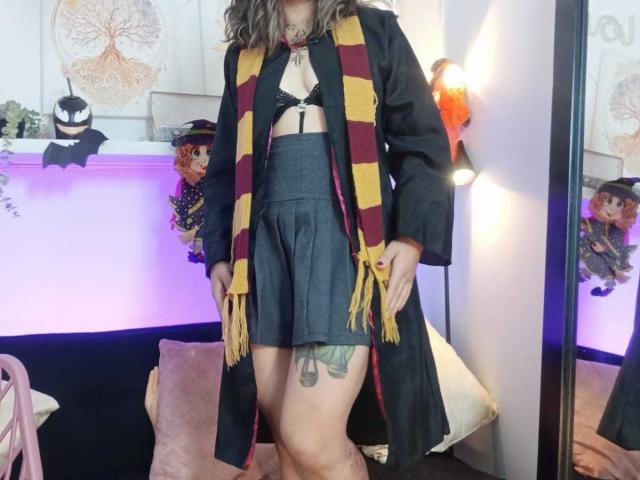 ArianaBlonde with wavy brown hair, wearing a black coat, yellow and red striped scarf, gray skirt, and black bra, showing a thigh tattoo and her ass.