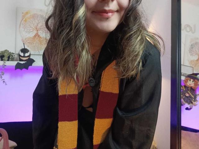 ArianaBlonde with wavy brown hair, black shirt, and red-yellow striped scarf, showing a pierced lip, and a black bra visible under the shirt. 