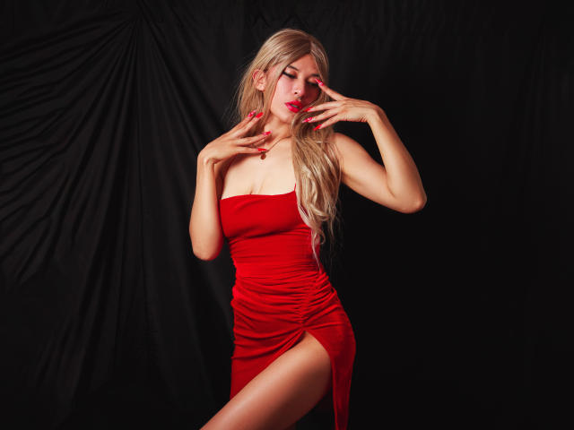 MartinaEvans with long blonde hair, red dress, and red nails, posing with hands near face, showing off her tits and a leg slit in the dress.