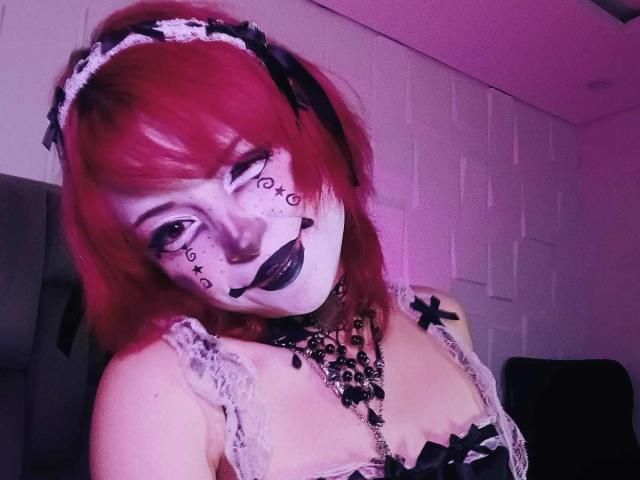 ViolettClay, con cabello rojo, lápiz labial negro, pintura blanca en la cara y "666" en las mejillas, vestida con una blusa de encaje, un collar negro y un lazo negro, en una habitación iluminada con luz púrpura..