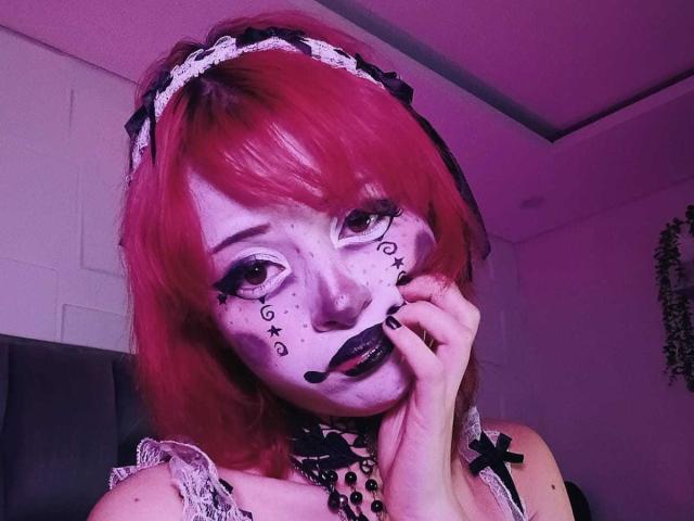 ViolettClay with red hair, white lace headband, black lipstick, and makeup dots on her face, touching her cheek, wearing a black and white lace top. Purple lighting.